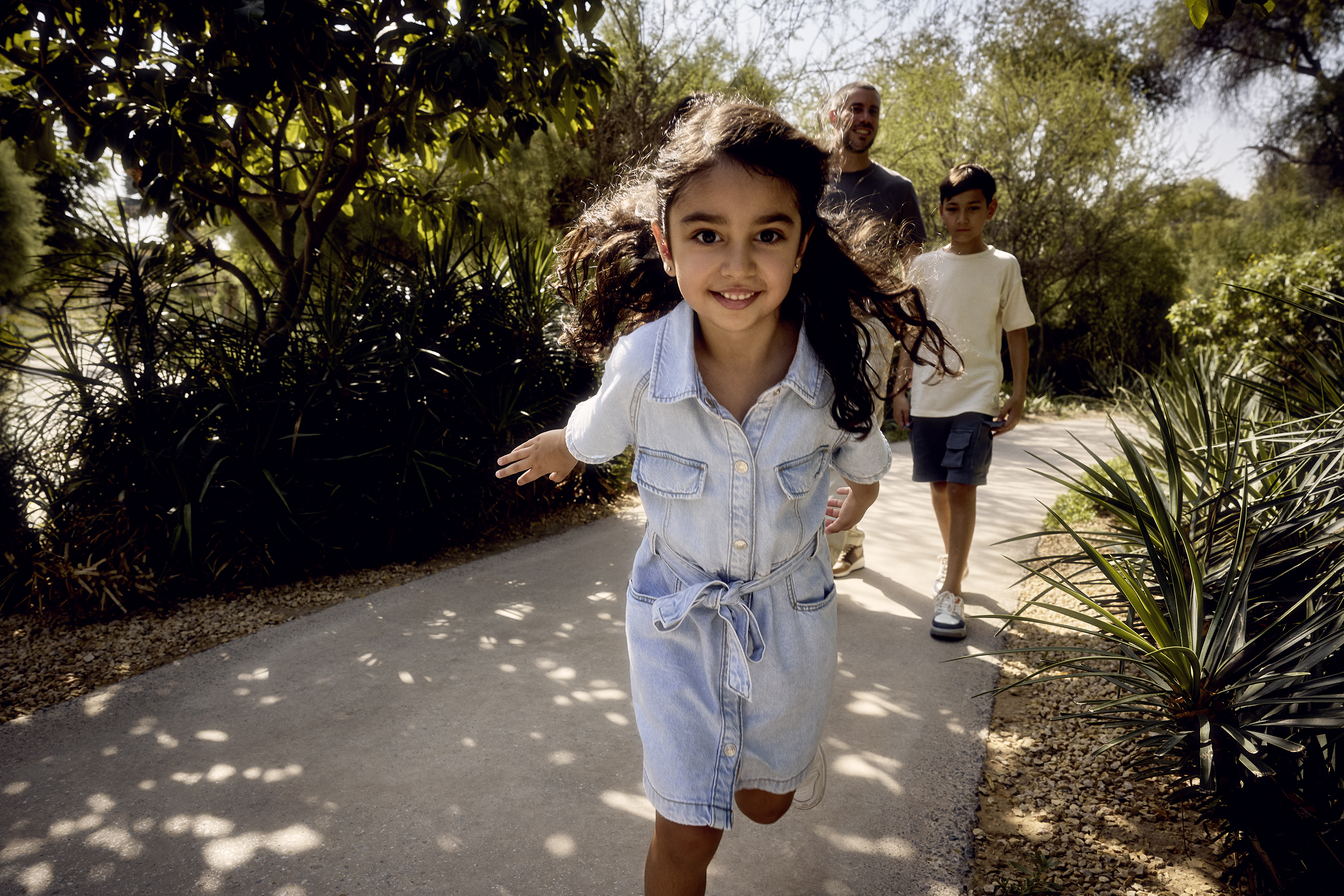 A girl runs toward the camera on a garden path, with a man and a boy walking behind her.