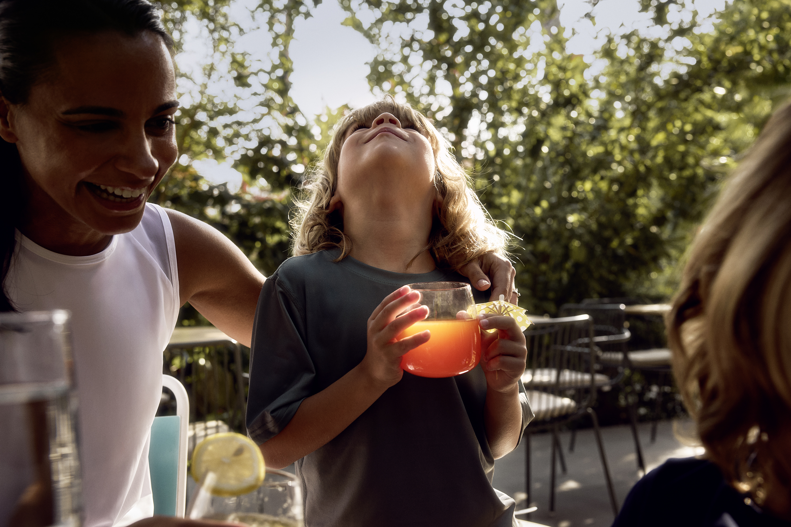 A woman and a child share drinks at an outdoor table under trees.